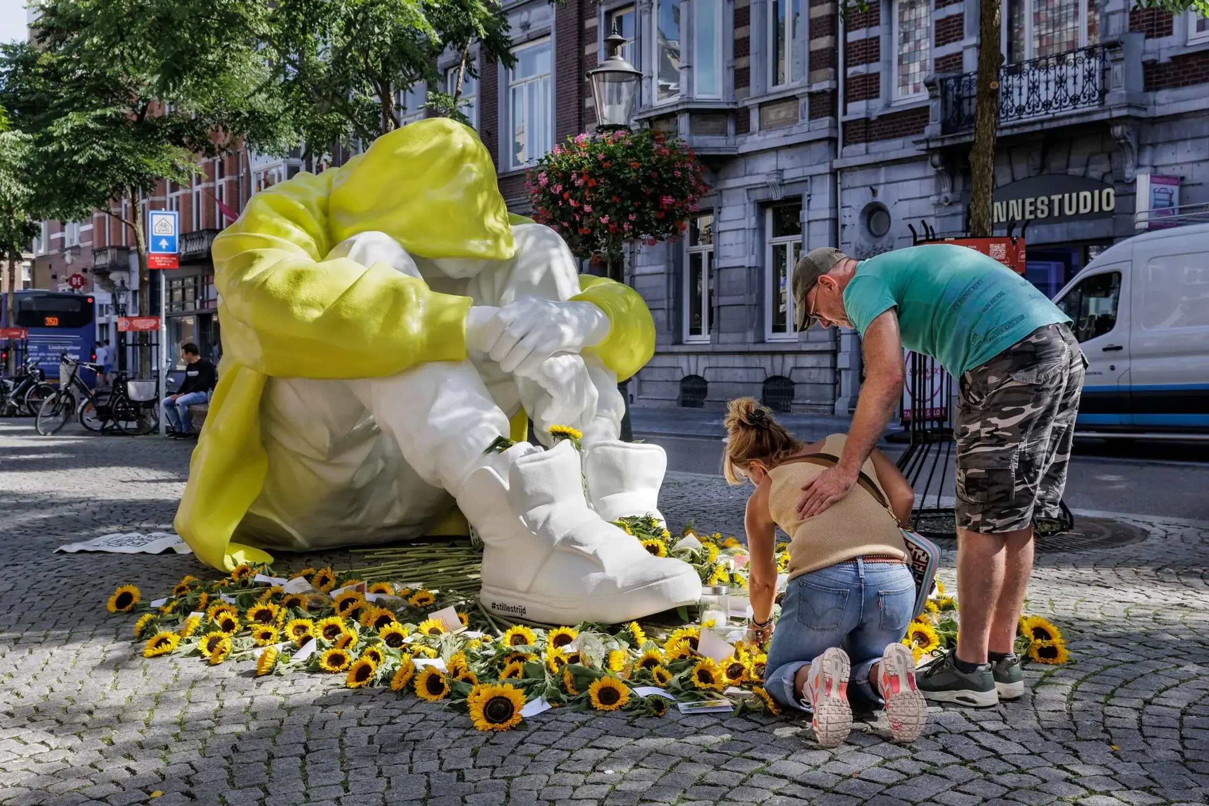 Een kunstwerk in Maastricht. Een reusachtige persoon zit teneeergeslagen op de grond. Mensen leggen er zonnebloemen bij